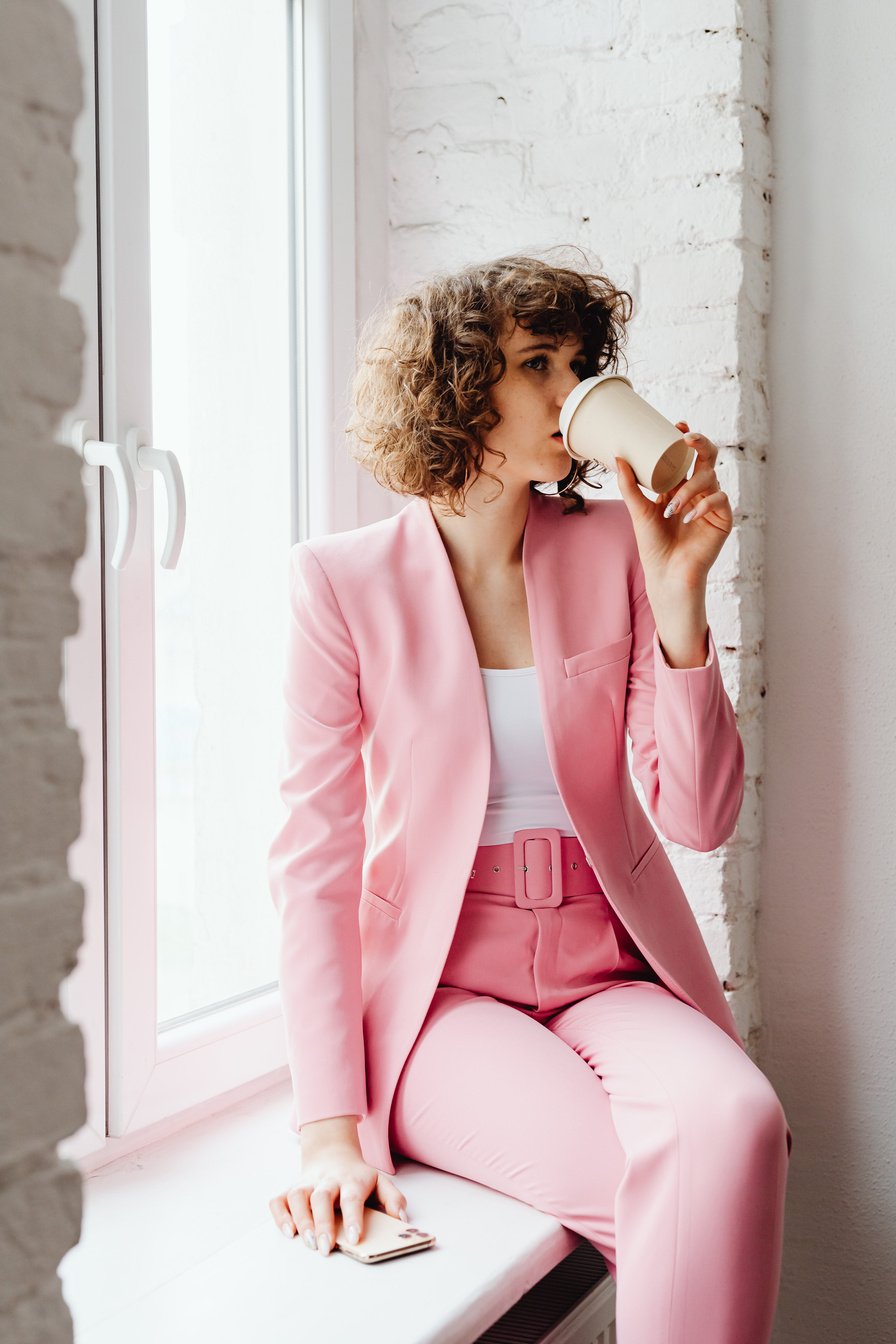 Woman in Pink Blazer Sitting Beside a Window Drinking Coffee